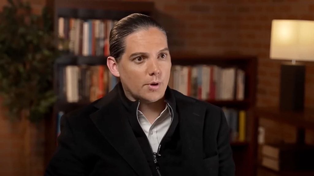 Adult person with slicked-back dark hair wearing a black jacket and white shirt, speaking in a warmly lit study with bookshelves and a lamp visible behind them.