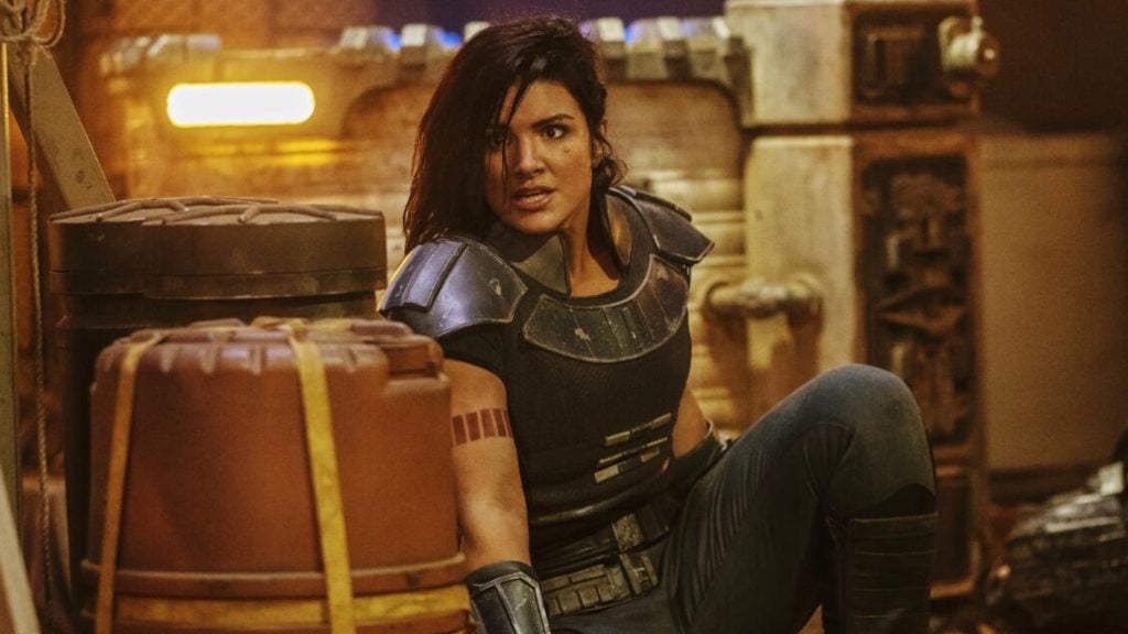 Gina Carano as Cara Dune, in black armor and a serious expression sitting on the floor next to large brown and black containers in a dimly lit industrial or sci-fi setting.