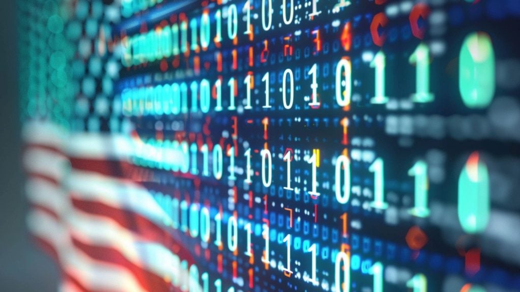 Blurred American flag in the background with an overlay of glowing binary digits (0s and 1s) and colorful data points across a digital display, evoking U.S. technology and cybersecurity.