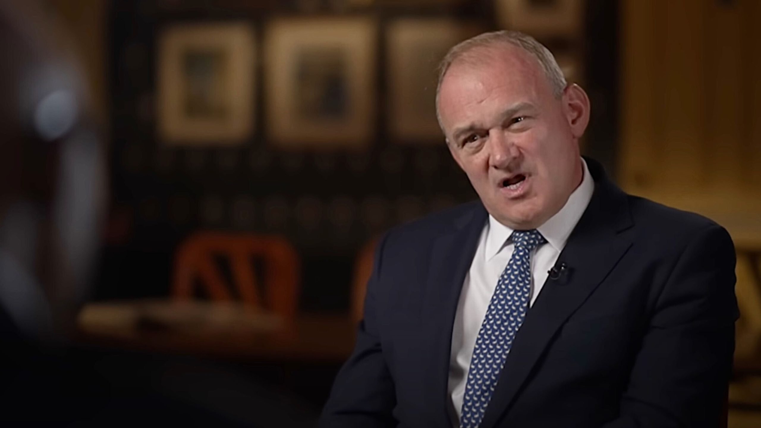 Davey in a dark suit and patterned blue tie speaking with a slightly open mouth during a filmed interview, photographed against a softly blurred interior background with framed pictures and wooden chairs.