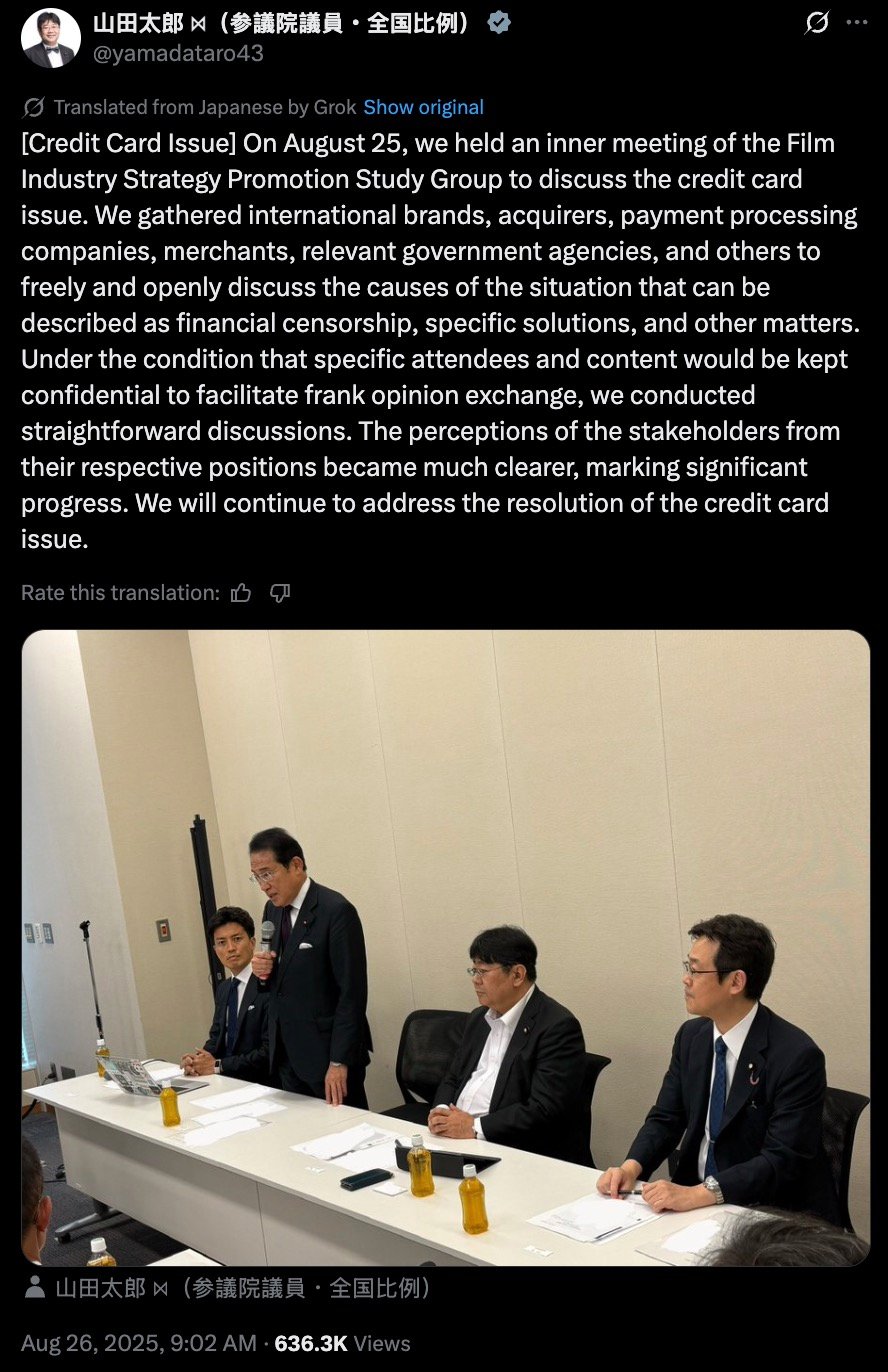 Four men in dark suits in a plain conference room around a long white table — one man stands speaking into a handheld microphone while the other three sit with papers, a laptop, and small bottled drinks in front of them.