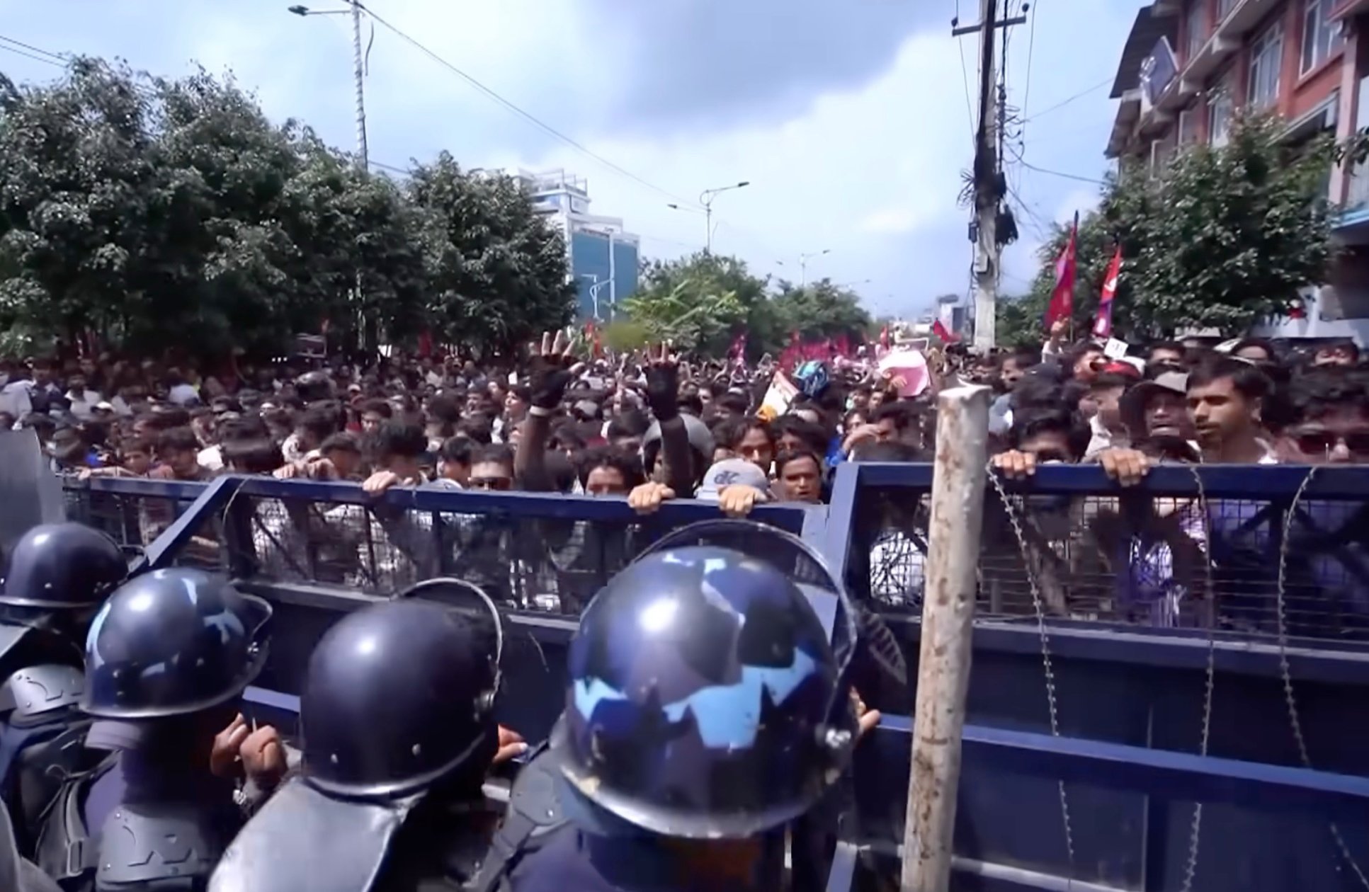 Crowd of protesters pressing against a blue metal barricade as helmeted riot police stand in the foreground on a city street with trees and buildings behind them.