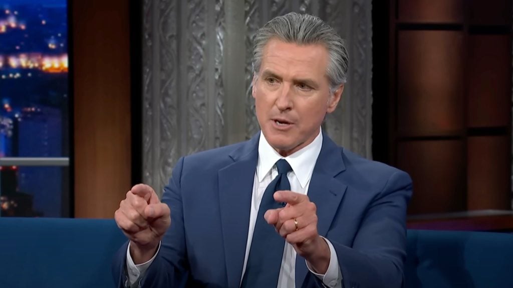 Governor Gavin Newsom with slicked-back gray hair in a blue suit, white shirt and dark tie sits on a talk-show set and points both index fingers forward while speaking, with a patterned metal backdrop and city lights visible through a window.
