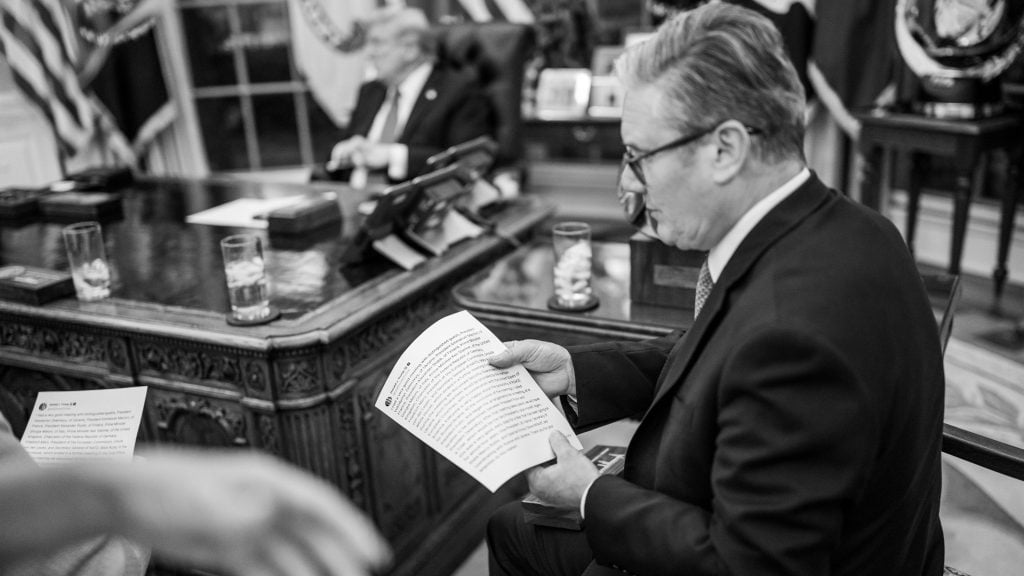 Black-and-white photo of Keir Starmer in a suit and glasses seated beside an ornate wooden desk, reading printed pages while a blurred figure sits behind the desk amid flags and glasses of water.