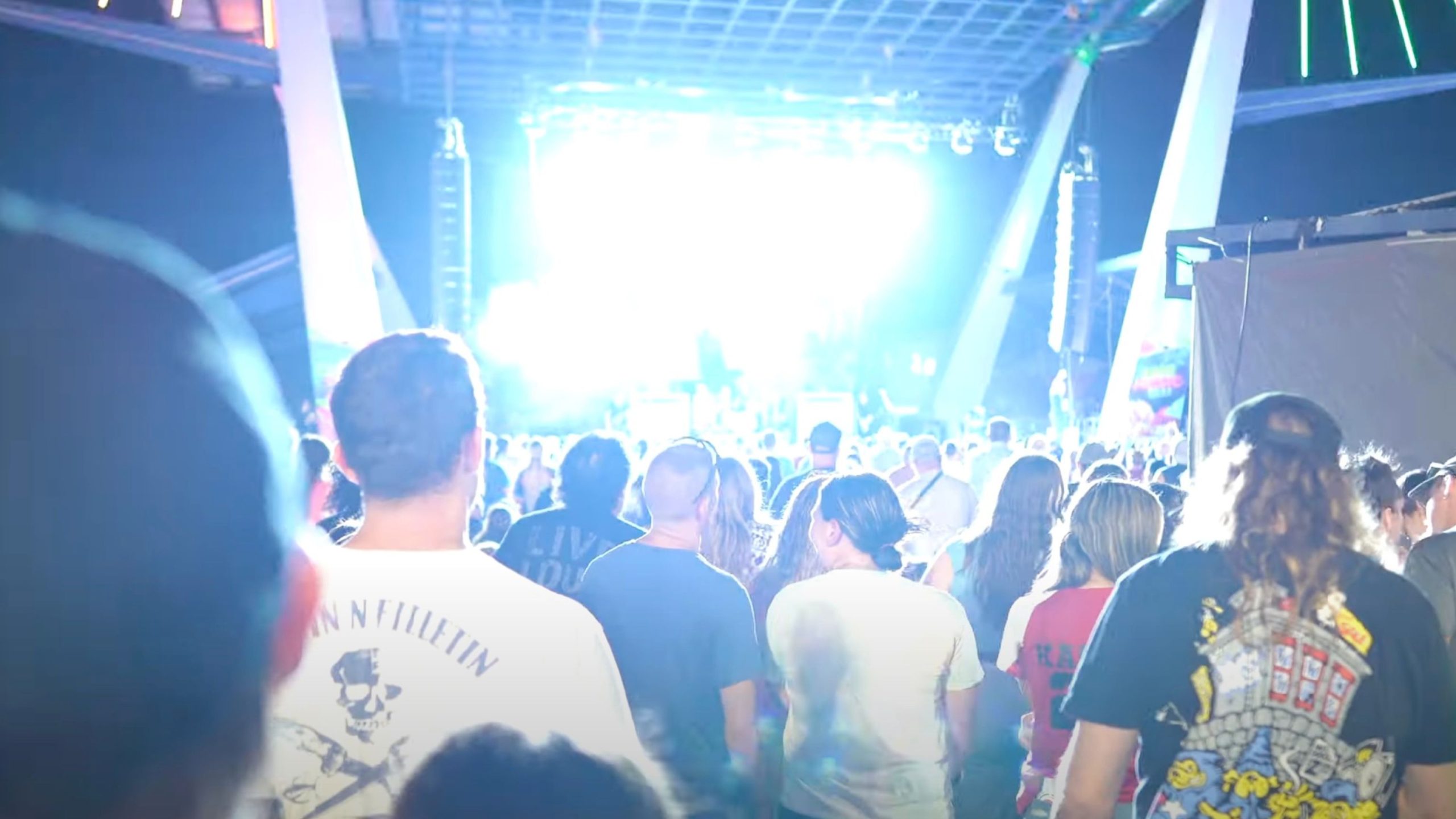 Crowd of people seen from behind watching an outdoor concert stage flooded with intense blue-white lights and large overhead rigging, their silhouettes and graphic T‑shirts visible in the foreground.