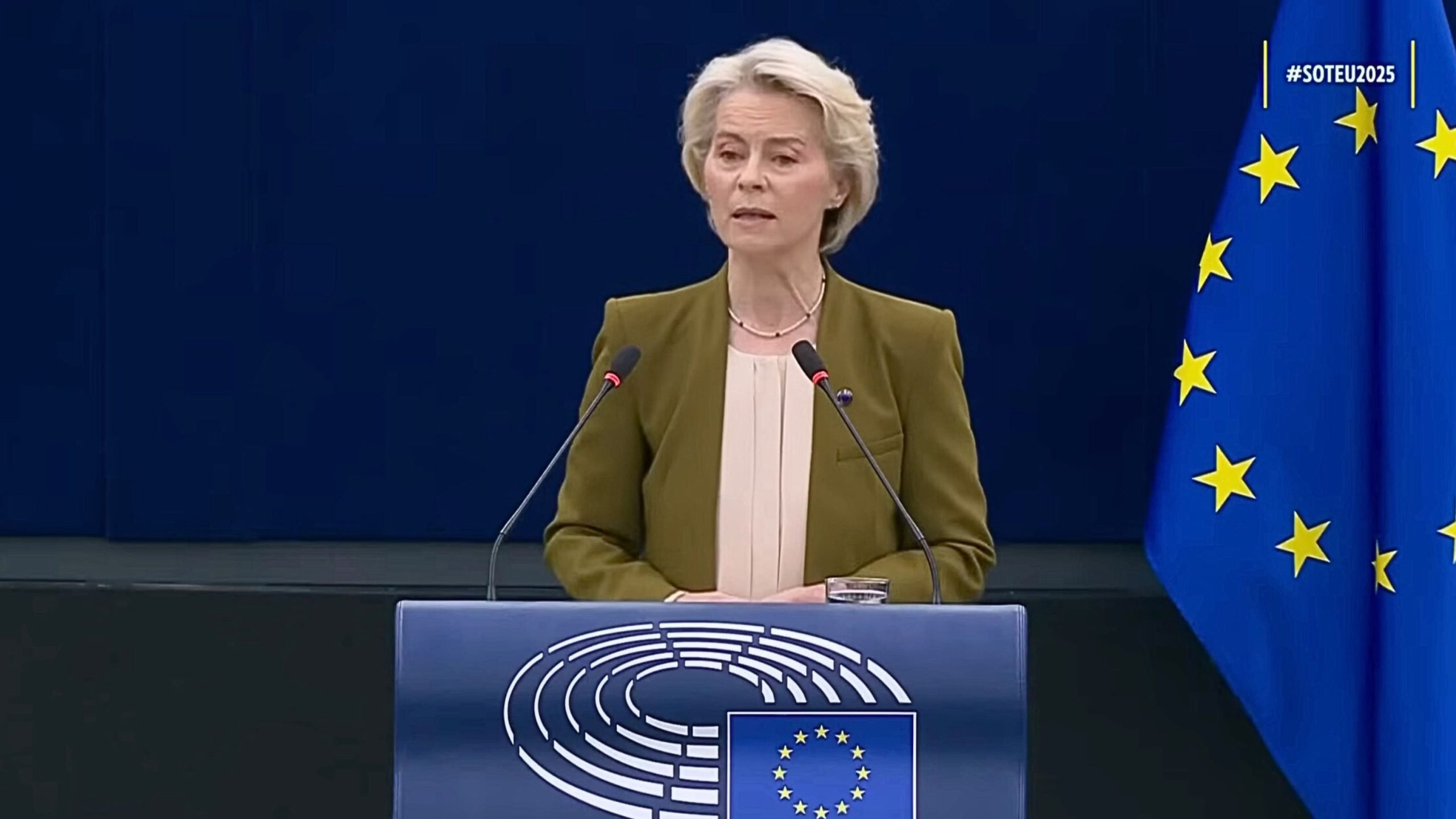 Von der Leyen in a green jacket speaking at a blue podium with the European Union emblem, two microphones in front of her and an EU flag to her right against a dark blue background.