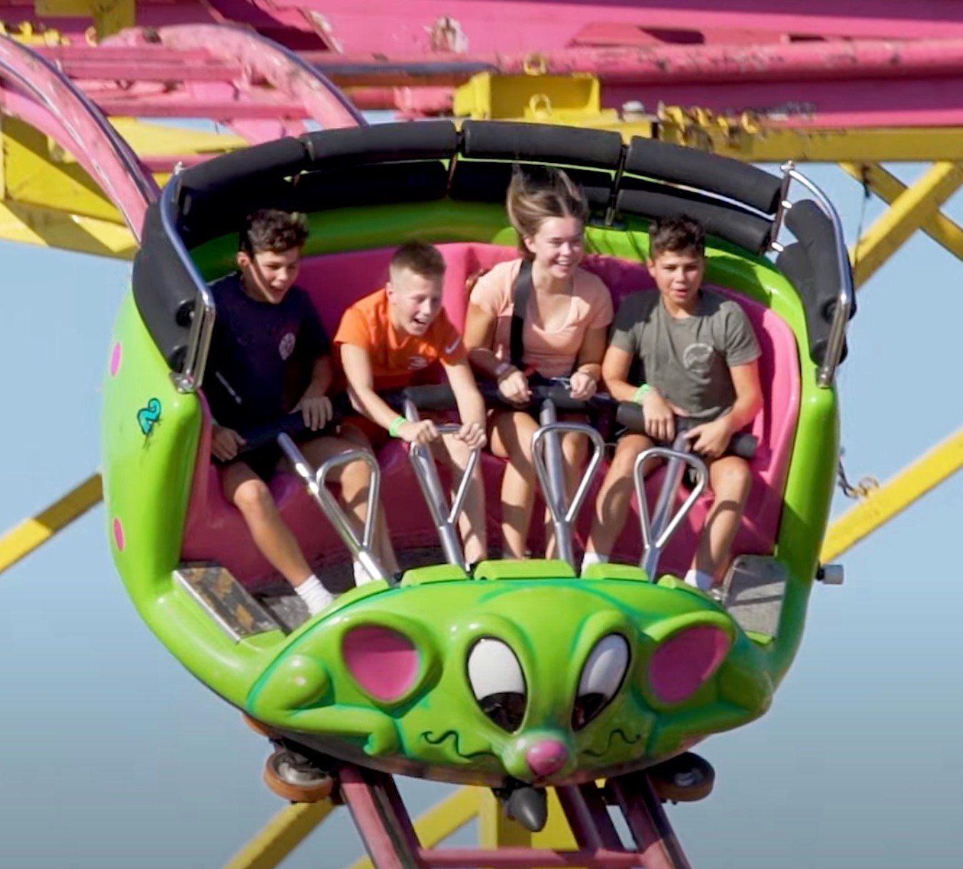 Four teenagers seated side-by-side in a bright green, mouse-faced roller coaster car at an amusement park, gripping safety bars and reacting with excited expressions against a clear blue sky.