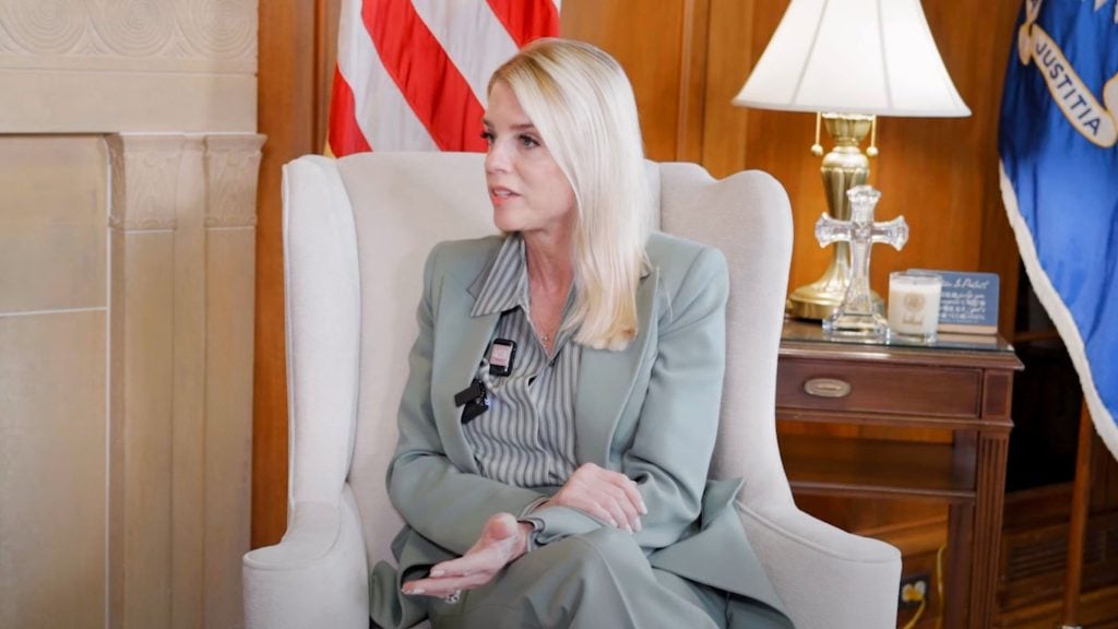 Pam Bondi in a light green suit sits in a beige wingback chair, gesturing as she speaks with an American flag and a wooden side table holding a lamp and a glass cross visible behind her.