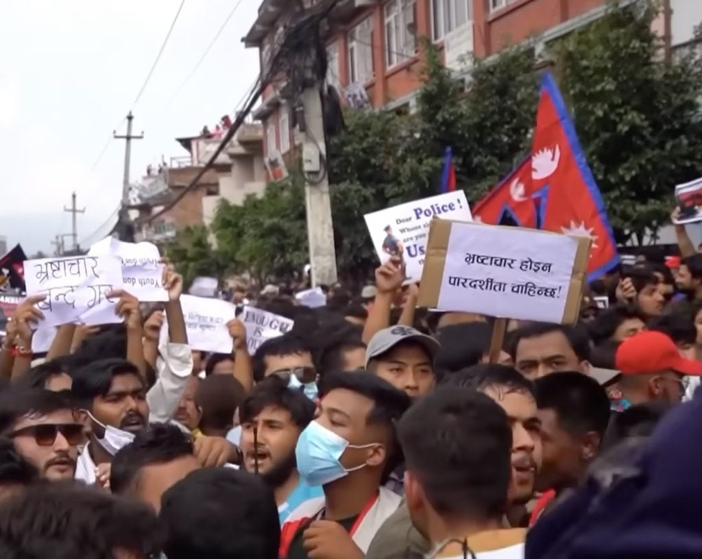 Dense crowd of protesters on a city street holding up placards—many with Nepali script—and waving red-and-blue Nepal flags, with a man in a blue surgical mask visible in the foreground and apartment buildings and utility poles lining the background.