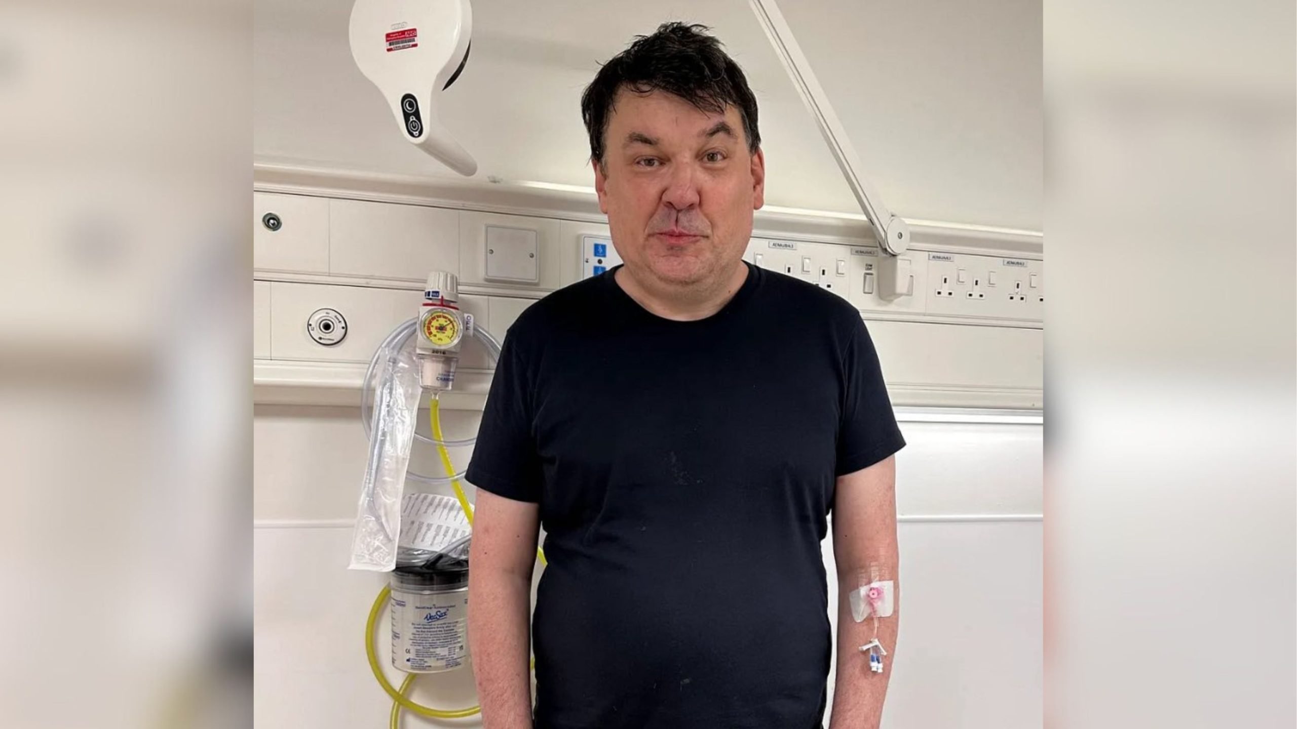 Graham Linehan in a black T-shirt standing in a hospital room with an IV cannula taped to his forearm and medical tubing and wall-mounted equipment visible behind him.