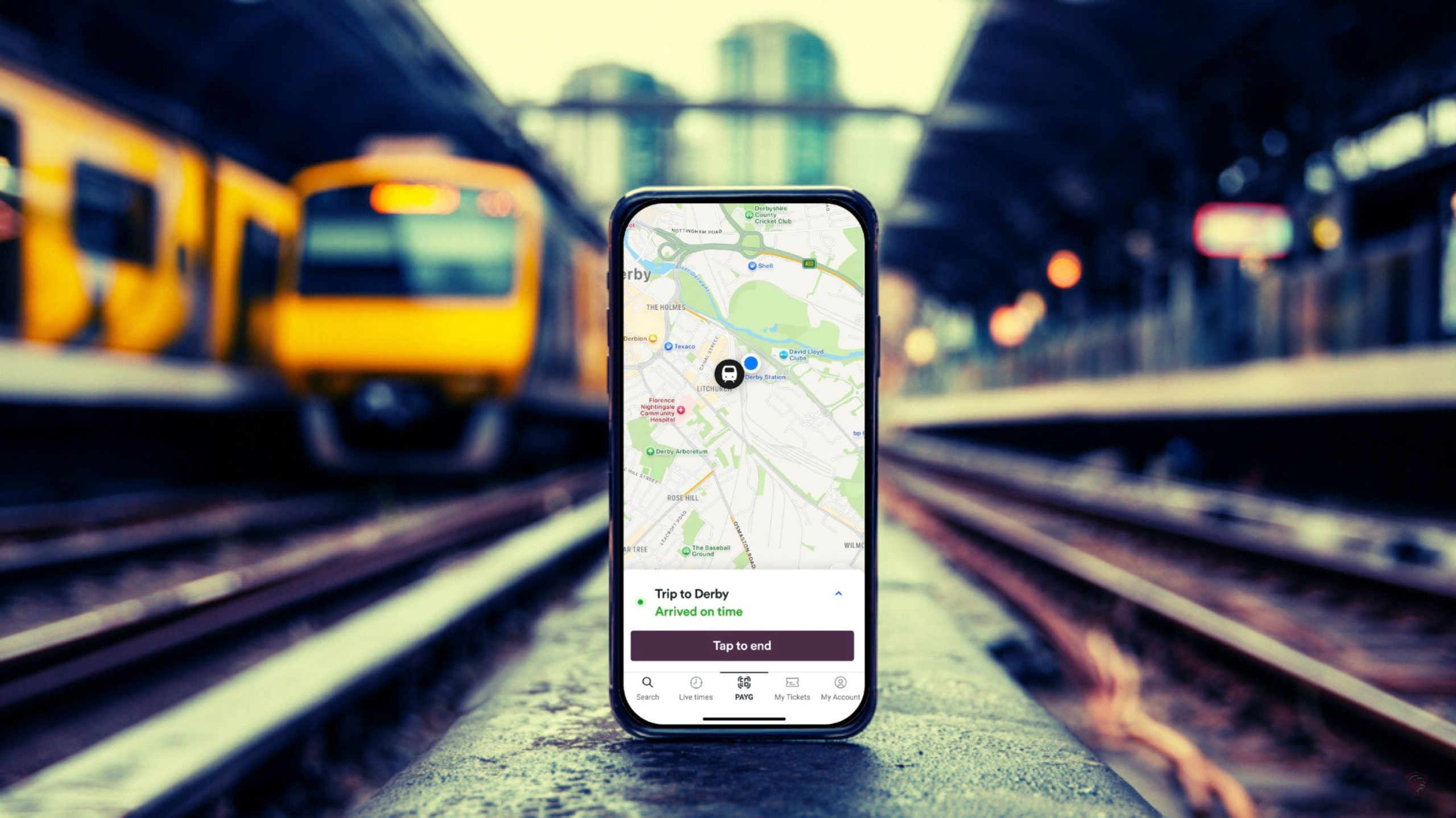 Smartphone standing upright on a train platform displaying a map and a notification reading 'Trip to Derby — Arrived on time', with blurred yellow train cars and railway tracks in the out-of-focus background.