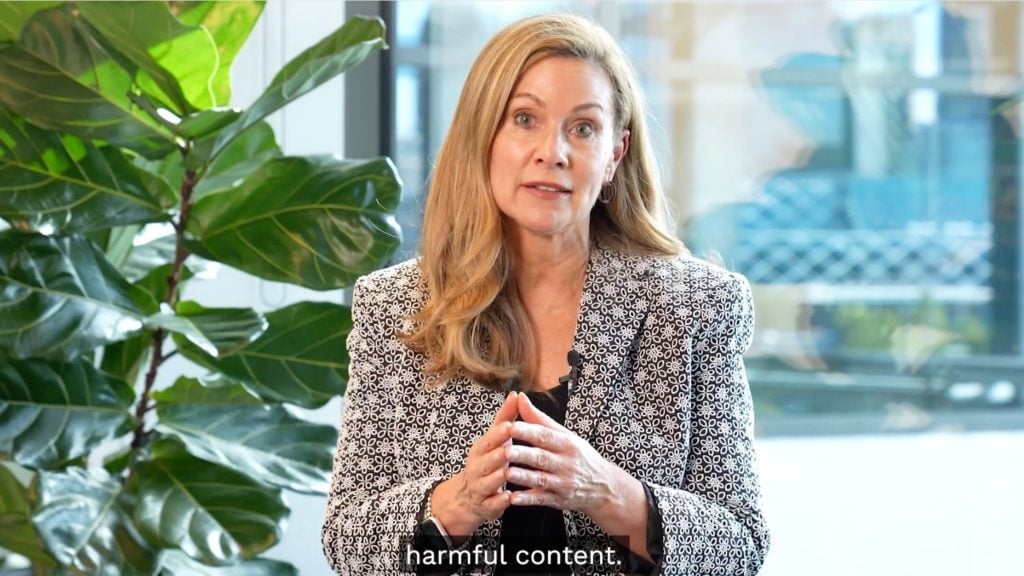 Grant with long blonde hair wearing a black-and-white patterned blazer speaks to camera with hands clasped, seated in a bright office next to a large leafy plant and the subtitle "harmful content" visible at the bottom.