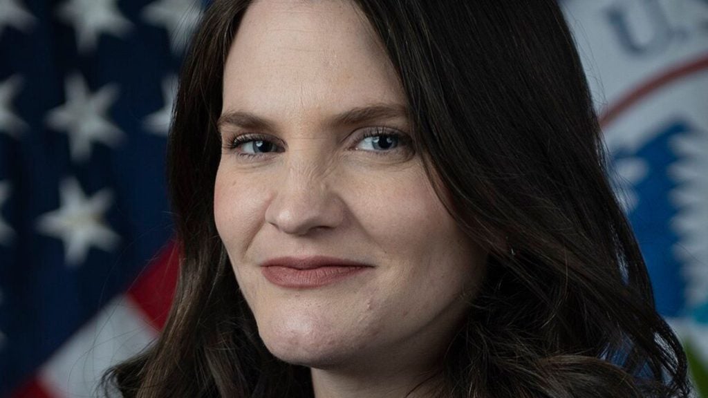 Close-up portrait of Nina with long dark hair and a subtle smile, with a blurred American flag and seal in the background.