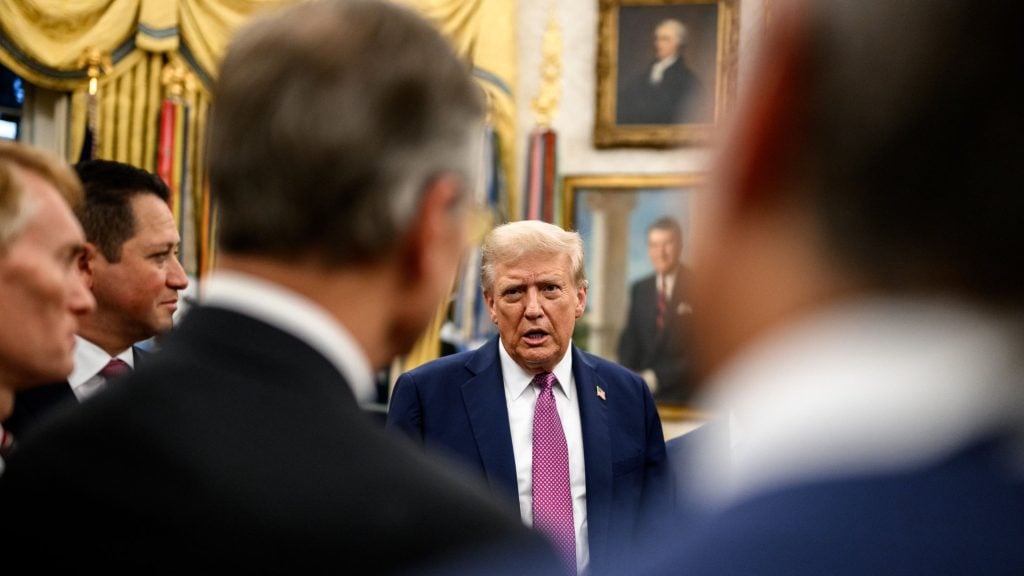 Trump wearing a navy suit, white shirt and pink patterned tie speaks in a formal room with gilded curtains and framed portraits while several blurred figures frame the foreground.