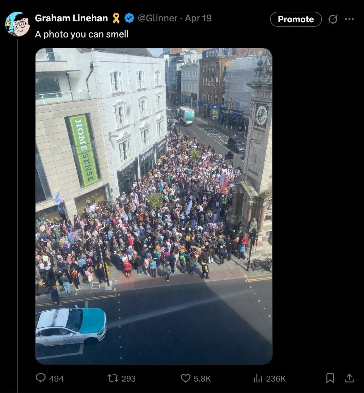 Screenshot of a social media post showing an aerial view of a dense crowd gathered on a city street corner outside a HomeSense store and a clock tower, many people holding blue-pink-white striped flags.