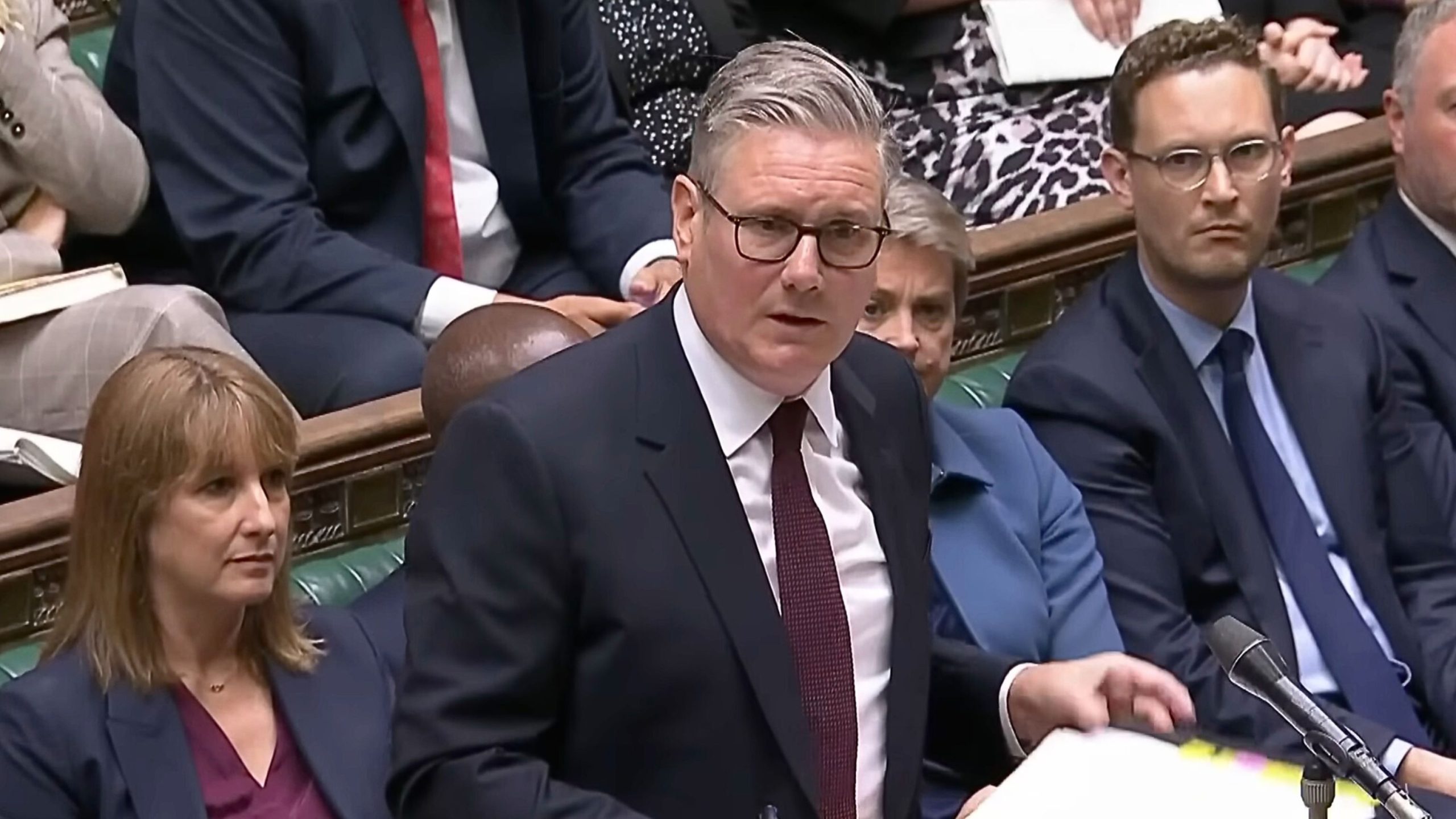 Starmer with gray hair and glasses in a dark suit and maroon tie speaking at a microphone in a wood-paneled parliamentary chamber, holding papers with colleagues seated behind him on green benches.