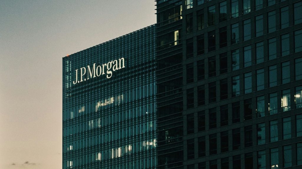 Dusk view of a J.P. Morgan logo illuminated on the glass facade of a high-rise office building, next to a darker grid of windows with a few interior lights visible.