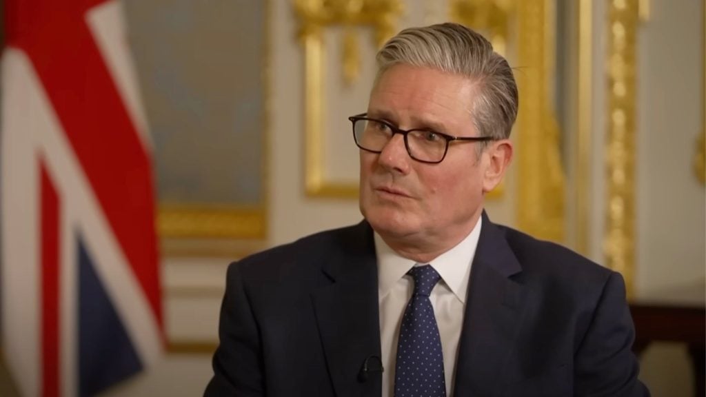 Starmer with gray hair and glasses wearing a dark suit and polka-dot tie, seated in an ornate room with a blurred British flag in the background.
