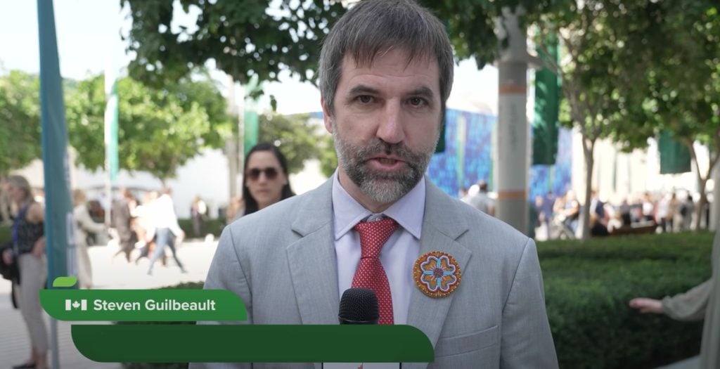 Guilbeault with salt-and-pepper beard wearing a light gray suit, red polka-dot tie and beaded floral pin speaks into a microphone outdoors with blurred trees and people in the background.