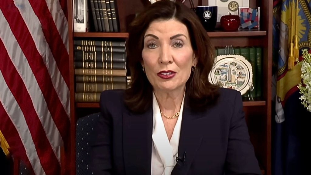 Hochul with shoulder-length dark hair wearing a navy blazer and white blouse speaking from an office, seated before a bookshelf filled with bound volumes, a decorative plate, and two flags including the U.S. flag.