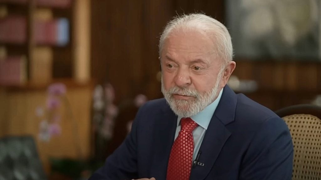 Lula da Silva with white hair and a trimmed beard wearing a navy suit, light blue shirt and red patterned tie, seated in a wood-paneled room and speaking with a thoughtful expression.