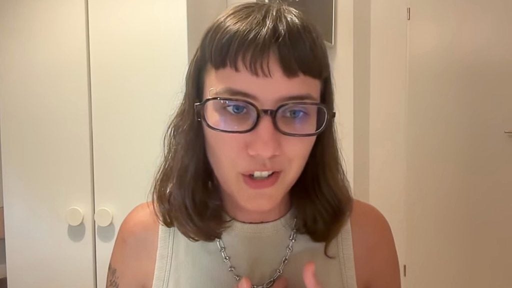 Isabella Cêpa with shoulder-length brown hair, short blunt bangs and rectangular glasses speaking on camera, wearing a sleeveless beige top and chain necklace in front of a white wardrobe.