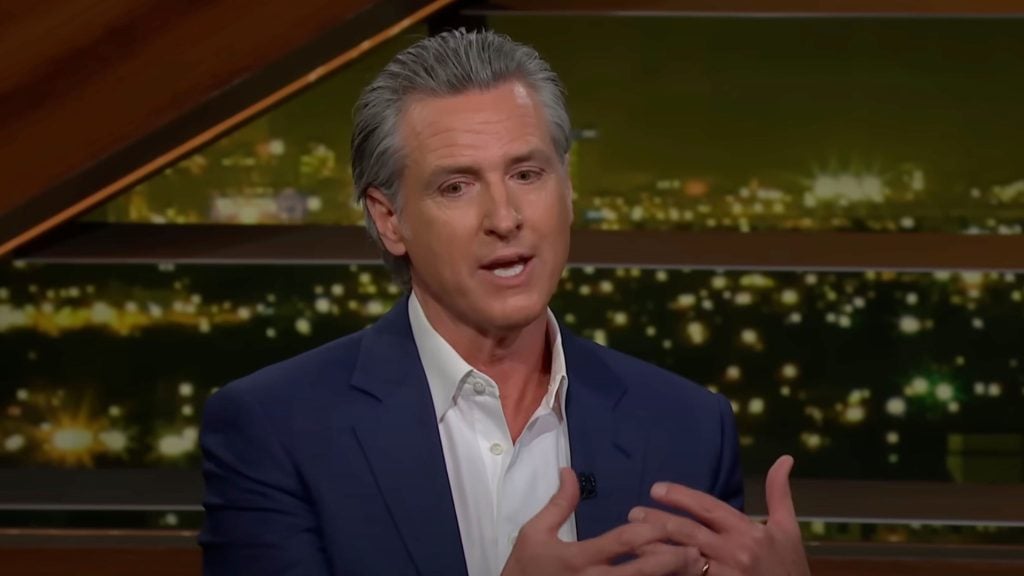 Newsom with salt-and-pepper hair in a navy blazer and white shirt speaking while gesturing with his hands, seated against a blurred nighttime city lights backdrop.