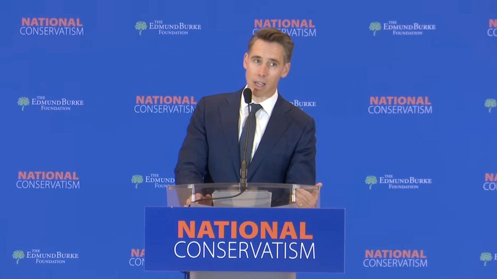 Josh Hawley speaks at a clear lectern with a microphone in front of a blue backdrop patterned with "National Conservatism" and "The Edmund Burke Foundation" logos.