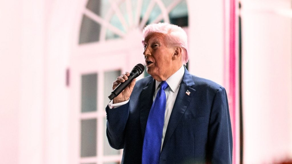 Trump light-colored hair in a dark blue suit and bright blue tie speaks into a handheld microphone, wearing a small U.S. flag lapel pin, standing in front of a light-colored building with paned windows.