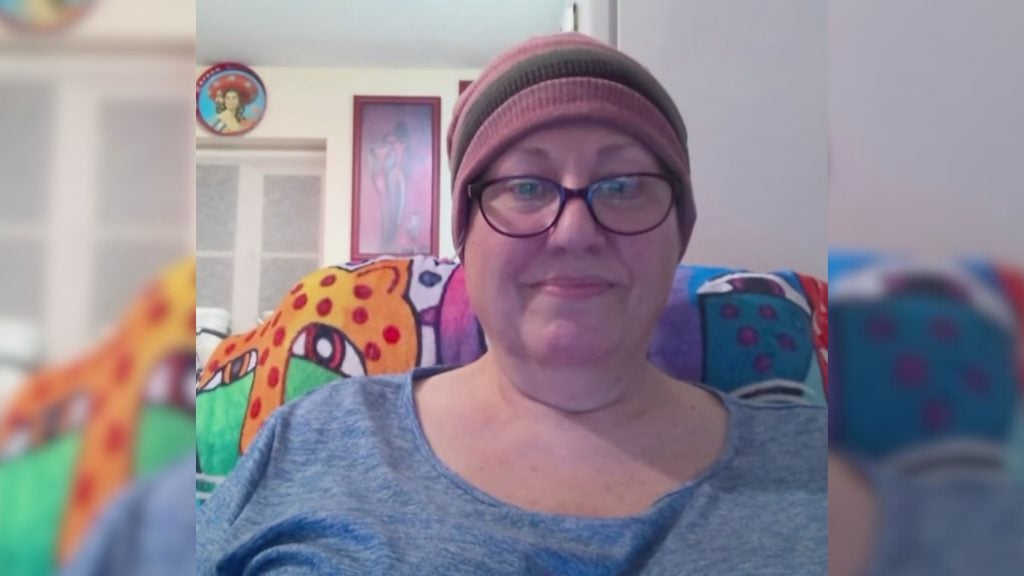 Anderson wearing glasses and a pink-and-brown striped beanie, smiling slightly while seated against a colorful patterned blanket with framed artwork and a doorway visible behind them.