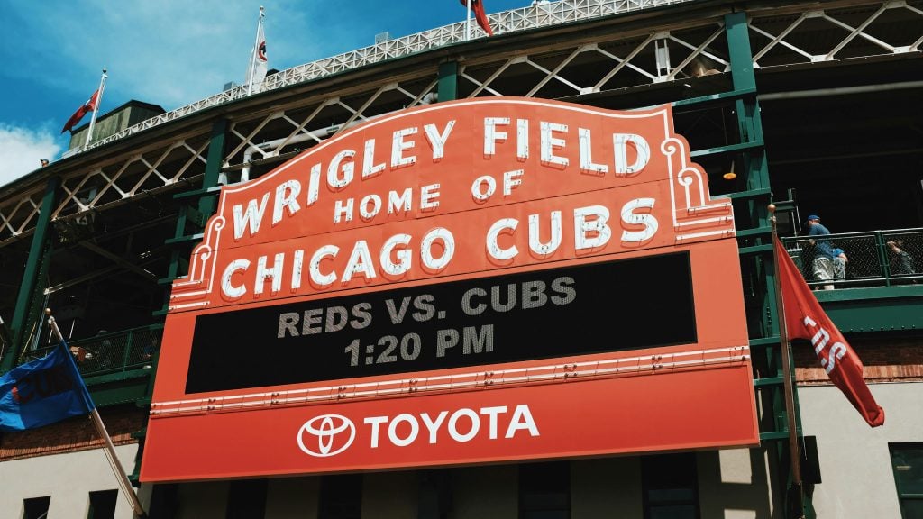 Wrigley Field's red marquee reading Wrigley Field Home of Chicago Cubs with an electronic board announcing Reds vs. Cubs 1:20 PM and a Toyota logo against the stadium's green steel facade and flags.