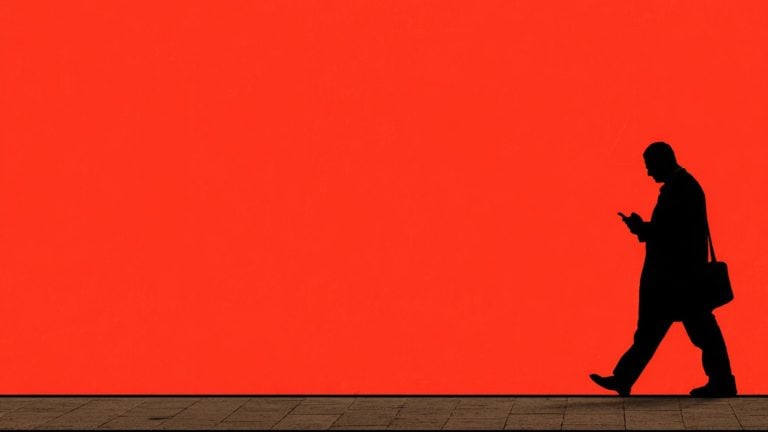 Black silhouette of a person in a coat walking to the right while looking at a smartphone and carrying a shoulder bag, set against a large flat red wall above a tiled sidewalk.