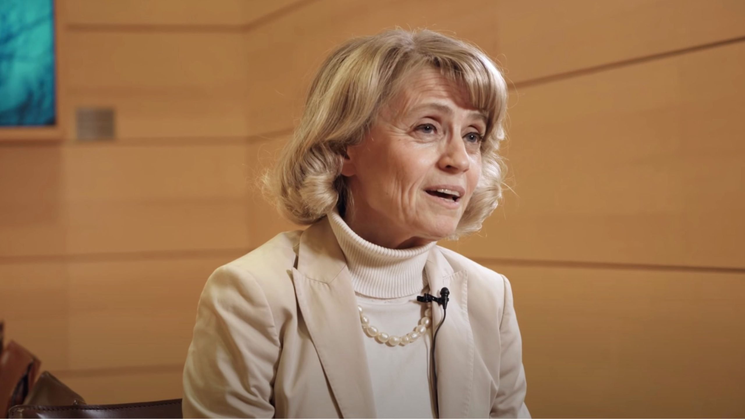 Older woman with short curled blonde-gray hair wearing a cream blazer, white turtleneck and a pearl necklace, speaking with a lapel microphone against a wood-paneled background.