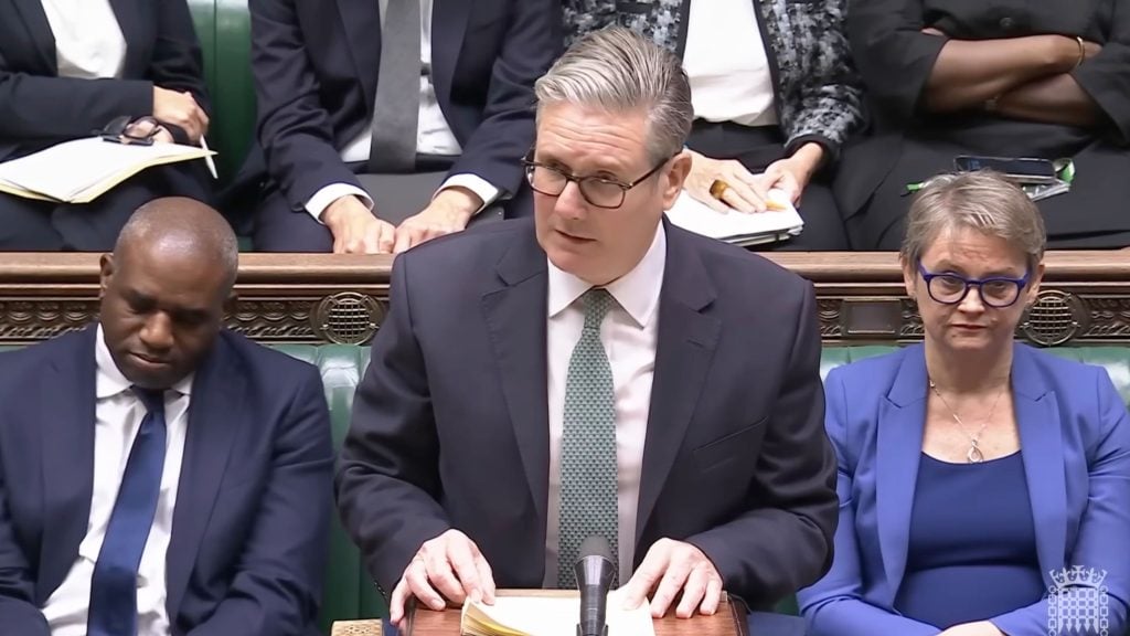 Starmer in a dark suit and green patterned tie stands at a wooden lectern with a microphone and papers, speaking in a parliamentary-style chamber while colleagues sit on green leather benches—one man with his head bowed on the left and a woman in a blue jacket and round glasses on the right.