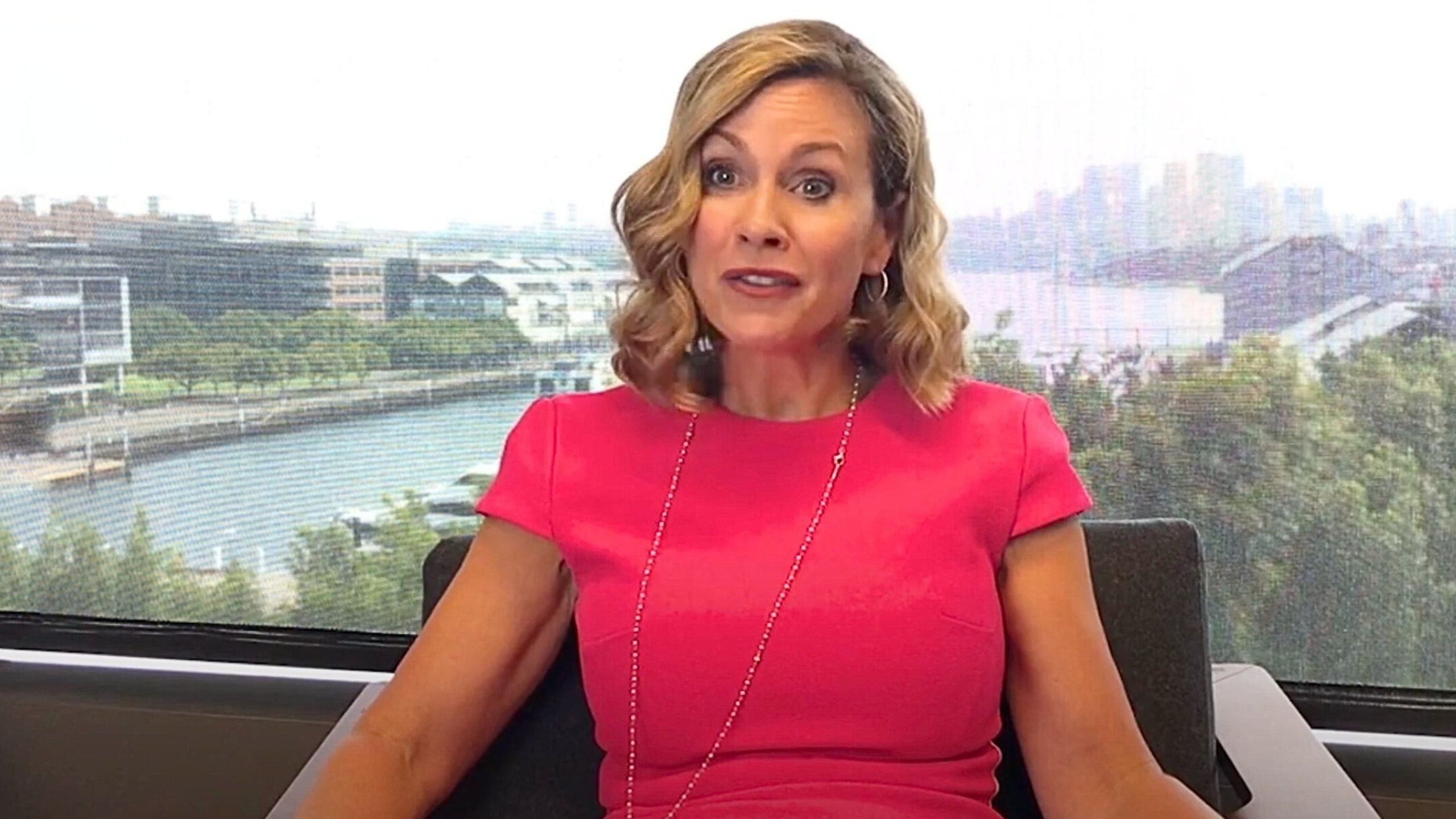 Inman Grant with shoulder-length wavy blonde hair wearing a bright pink dress and long beaded necklace sits in an office chair in front of a large window overlooking a river or harbor and a hazy city skyline, her face showing an engaged, animated expression.