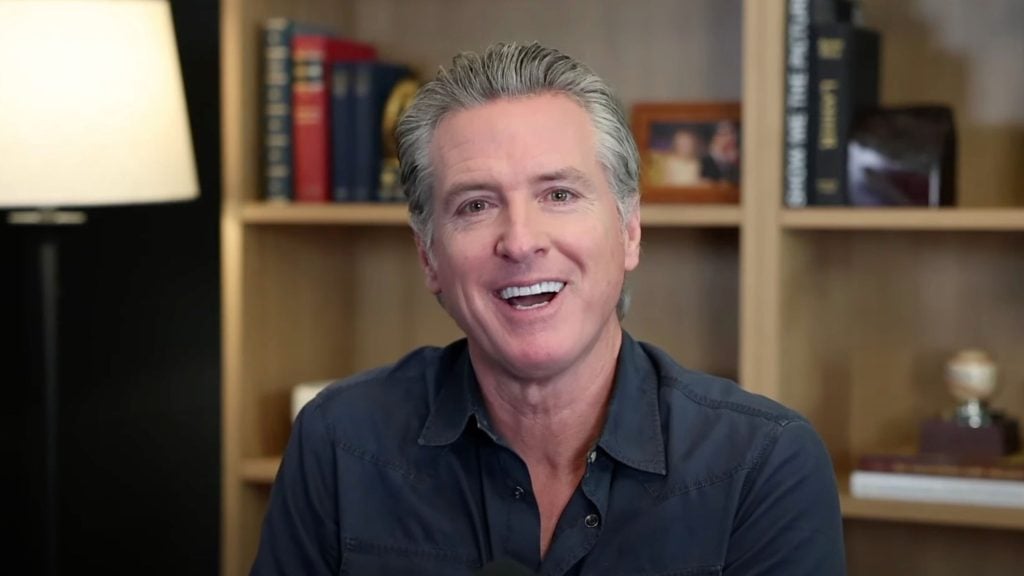 Newsom with slicked-back gray hair smiling at the camera, wearing a dark button-up shirt and seated in front of wooden bookshelves with books, a small framed photo and a lamp visible to the left.