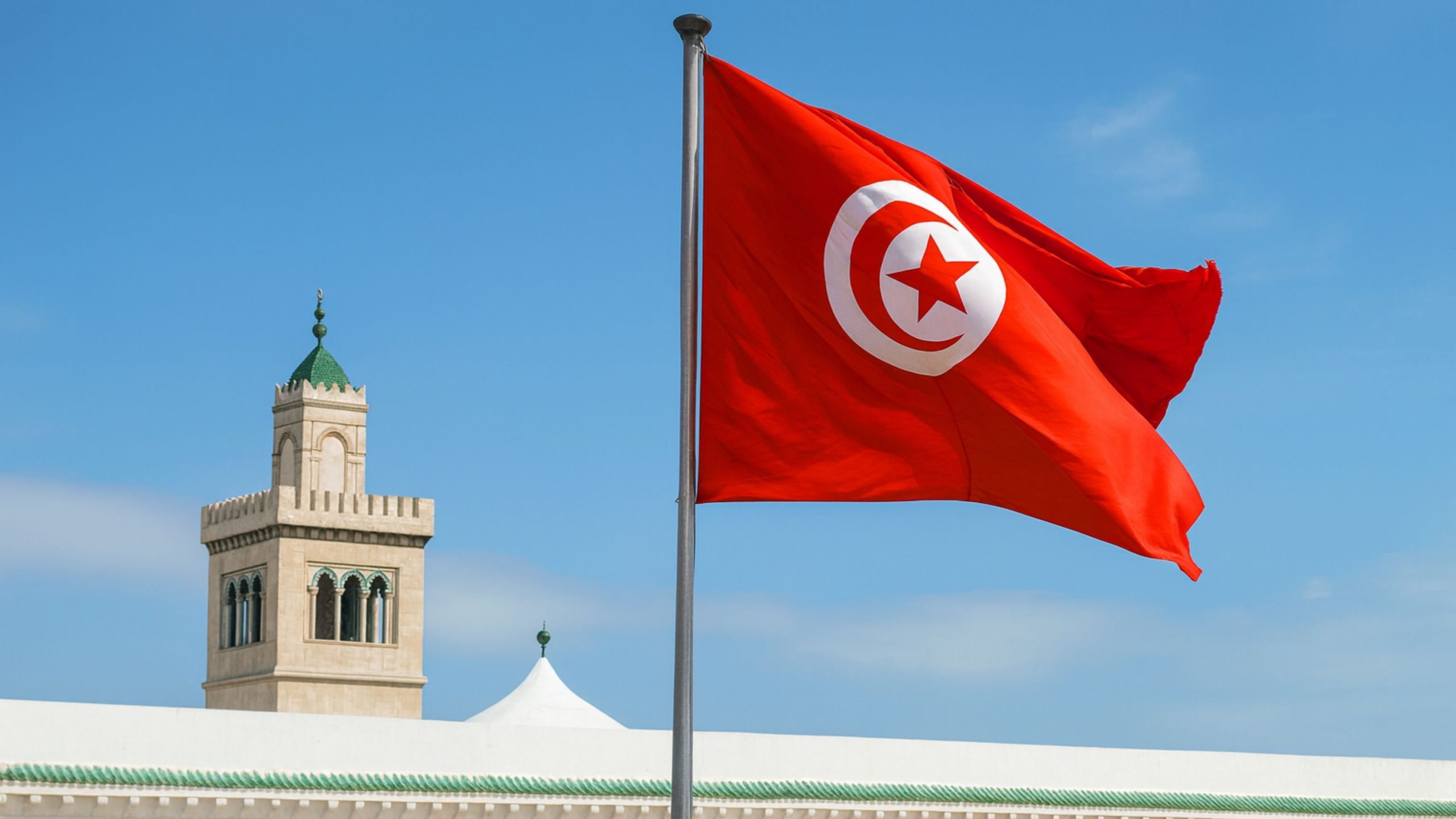 Red Tunisian flag bearing a white circle with a red crescent and five-pointed star, waving on a pole against a clear blue sky beside a beige mosque minaret topped with a green-tiled roof.