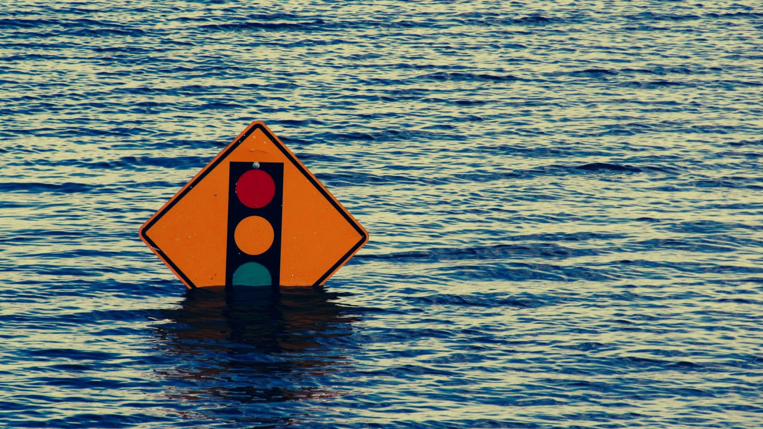 Diamond-shaped orange road sign depicting a vertical traffic light with red, yellow and green circles, partially submerged in rippling floodwater.
