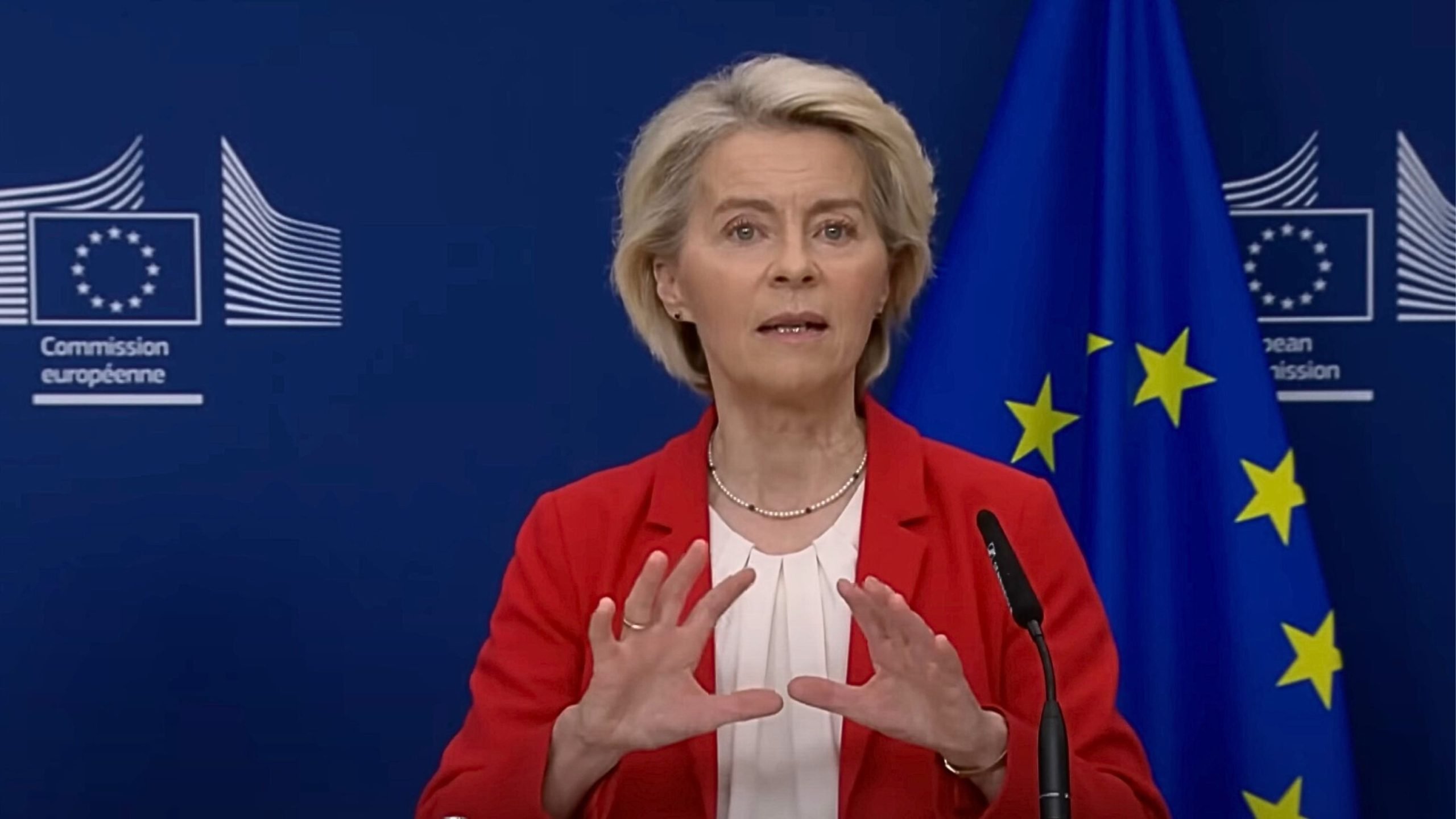 Von de Leyen with blond hair wearing a red blazer and white blouse speaking at a podium with hands raised, EU flag and European Commission logos on a dark blue backdrop