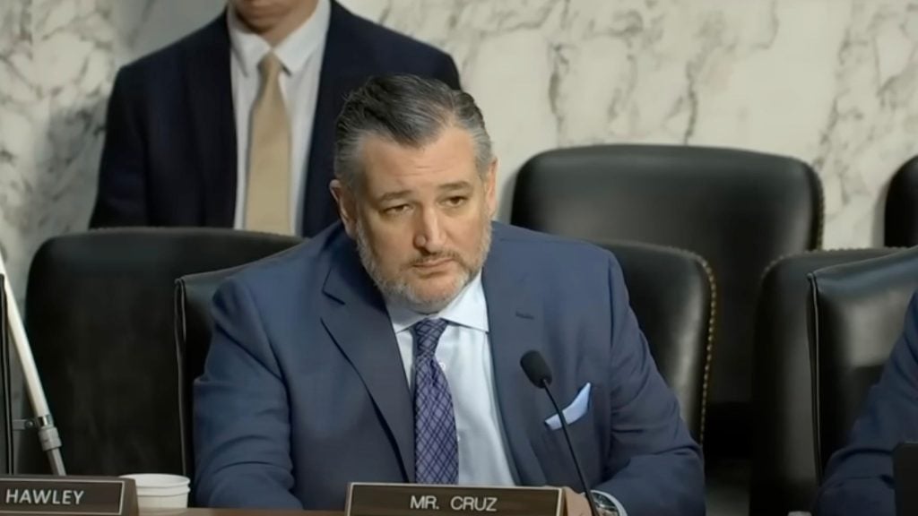 Ted Cruz in a blue suit and patterned tie seated at a formal hearing desk, speaking into a microphone with leather chairs and a marble wall background.