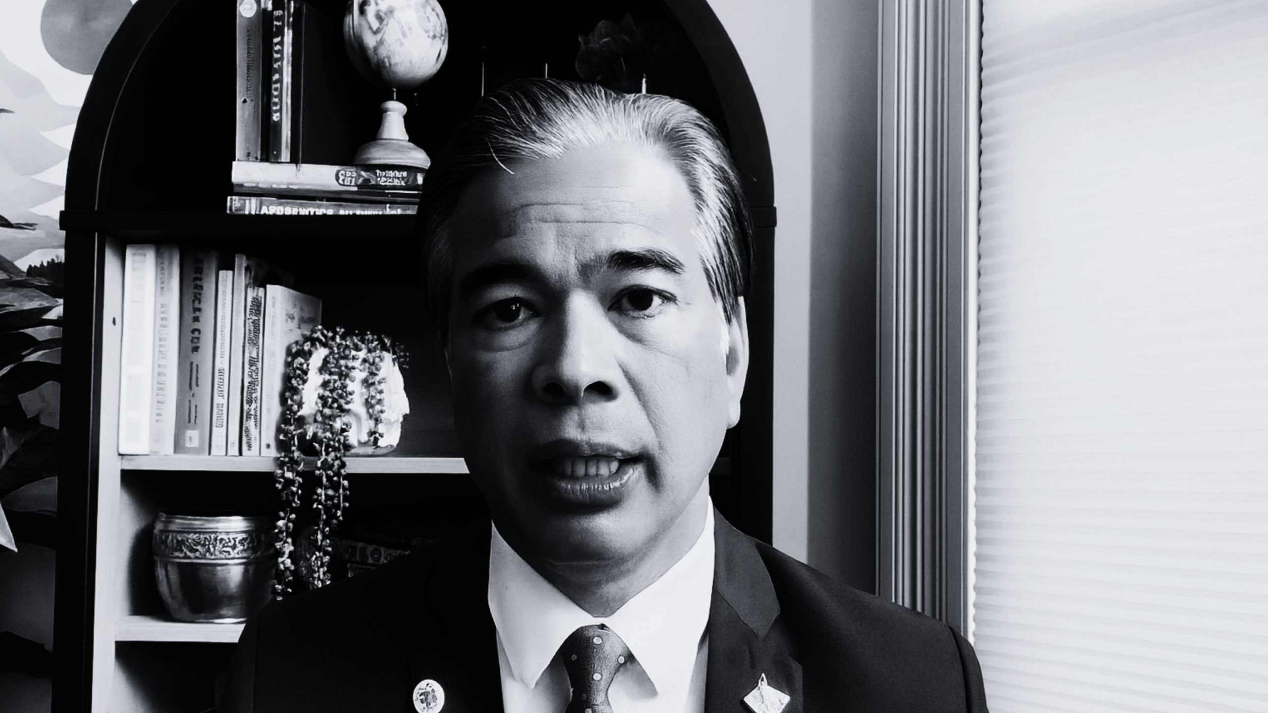 Bonta in a suit and tie speaking directly to the camera, framed against a bookshelf with books, decorative objects and a small globe on the left and window blinds on the right.