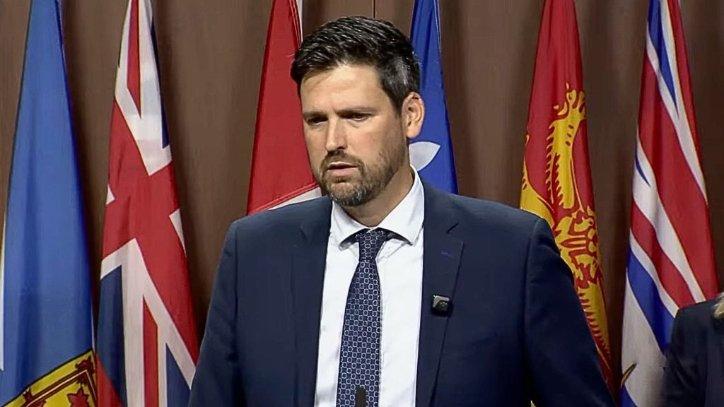 Sean Fraser in a dark suit and patterned tie speaking at a podium with a lapel microphone, standing in front of several red and blue provincial flags.