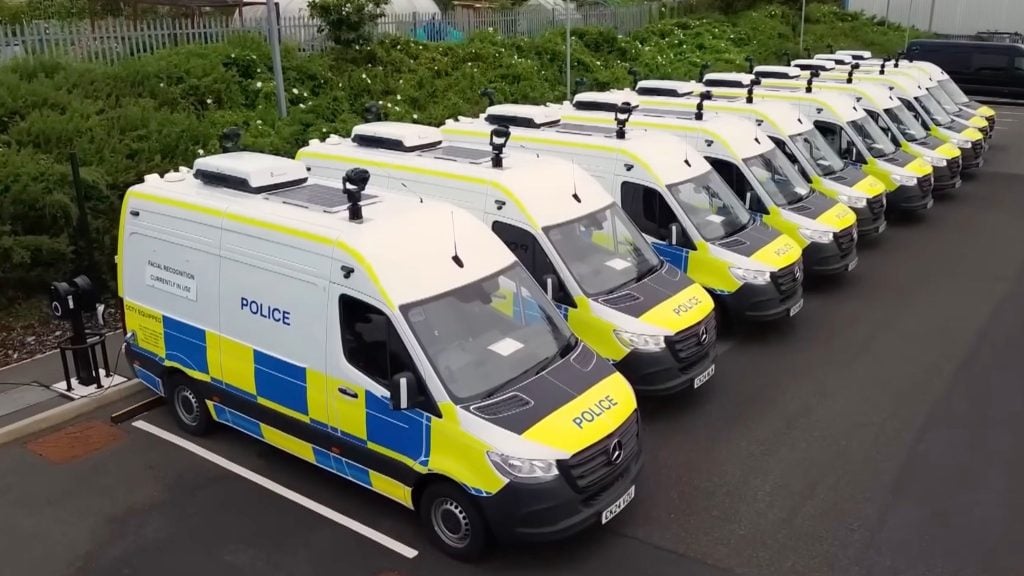 A line of white police vans with yellow high-visibility panels and blue chequered markings parked in a lot, each fitted with roof-mounted cameras and sensors and a notice on the nearest van mentioning facial recognition and CCTV equipment.