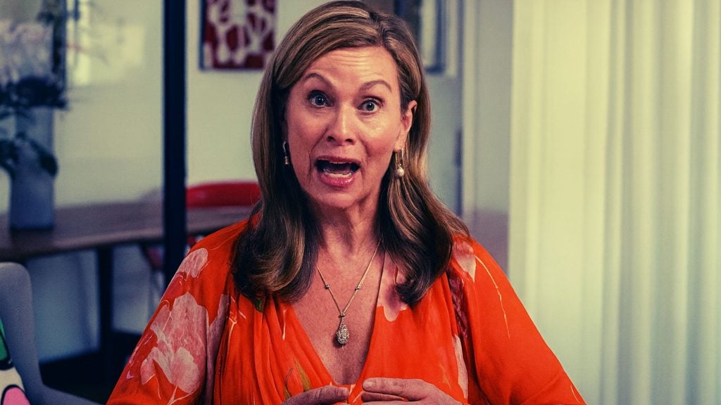 Inman Grant with shoulder-length light brown hair and dangling earrings, wearing a bright orange floral blouse and pendant necklace, seated indoors with an open-mouthed, expressive face as if speaking.
