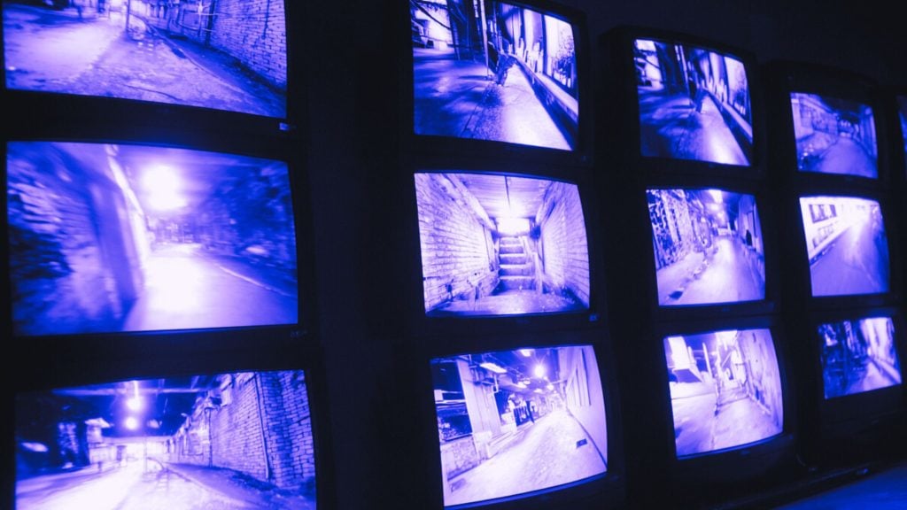 Wall of stacked monitors showing blue‑tinted surveillance footage of empty alleyways, staircases and corridors.