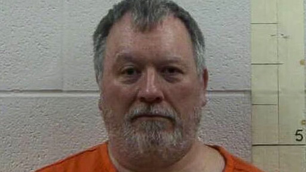 Frontal portrait of Larry Bushart with short graying hair and a trimmed gray beard, wearing an orange prison shirt against a textured cinderblock wall with height markings visible at the right.