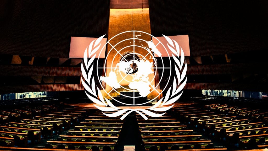 Empty, dimly lit assembly chamber with wooden tiered seating and a golden-lit central podium, overlaid by a large white United Nations emblem centered in the image.
