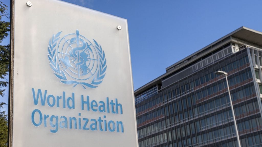 World Health Organization blue emblem and lettering on a freestanding sign in front of a modern glass office building against a clear blue sky.
