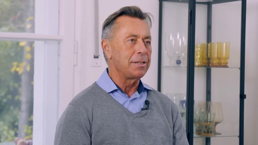 Norbert Bolz wearing a gray V-neck sweater over a blue striped collared shirt with a clip-on microphone, shown speaking indoors beside a glass display cabinet filled with yellow and clear glassware and a window with blurred greenery outside.