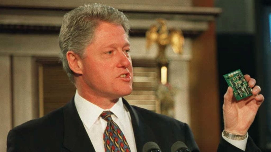 President Bill Clinton in a dark suit and patterned tie speaking at a podium while holding up a small green printed circuit board between his fingers.